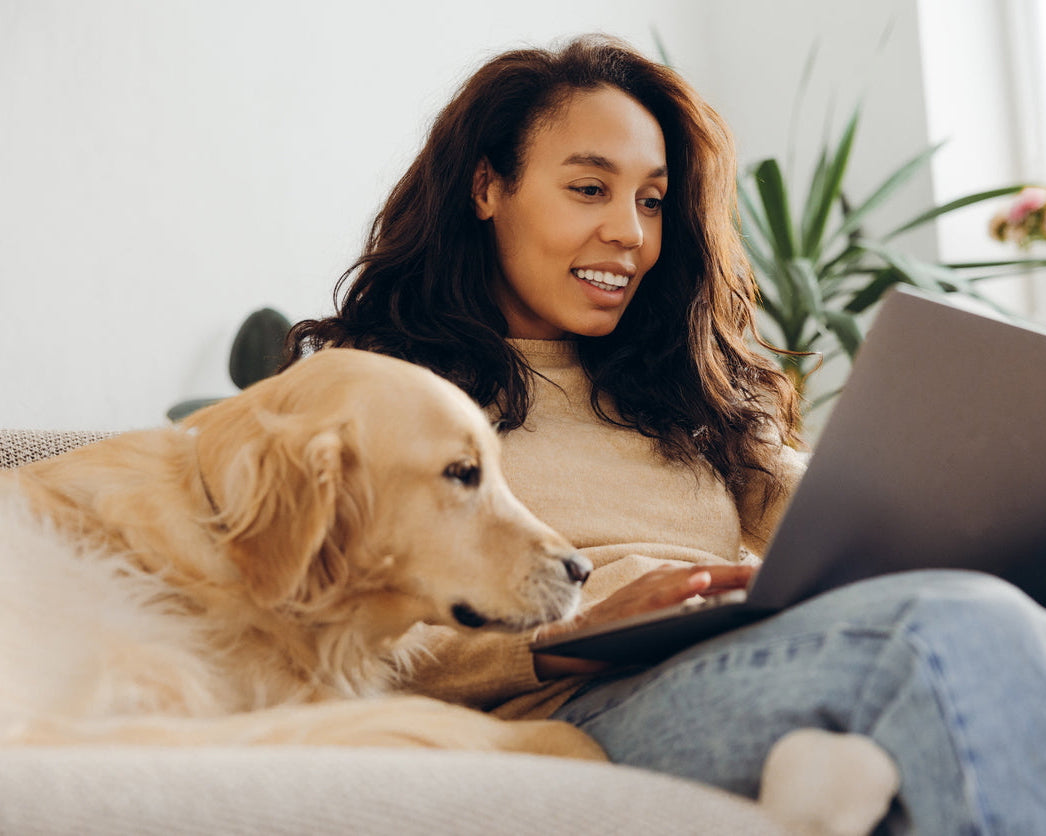 A woman sitting on a couch with a laptop, with a golden retriever dog resting beside her. The woman is wearing a beige top and blue jeans, and the scene is set in a bright, modern living room with a plant in the background.